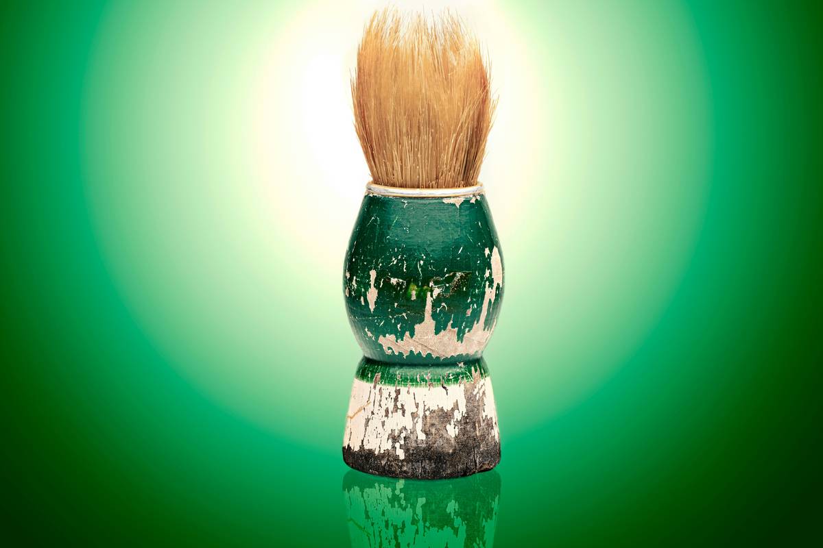 A hand holding a classic shaving brush swirling lather over a puck of traditional shaving soap on a wooden stand, highlighting traditional shaving soap economy benefits